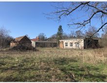 Ehemalige landwirtschaftliche Gebäude in einer offenen, grasbewachsenen Landschaft, bestehend aus einem Backsteinhaus mit Satteldach und einem flachen Gebäude aus Ziegeln und Beton, umgeben von kahlen Bäumen und Gestrüpp. Im Hintergrund sind weitere Gebäude und einige hohe Bäume zu sehen.