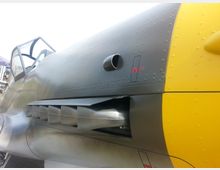 Nahaufnahme eines historischen Flugzeugs mit einer grau-gelben Lackierung, dargestellt ist ein Teil des Cockpits mit geöffneter Verglasung und der seitlichen Motorabdeckung, unter der Lamellen und Belüftungsöffnungen erkennbar sind. Nieten und Oberflächenstrukturen sind deutlich sichtbar.