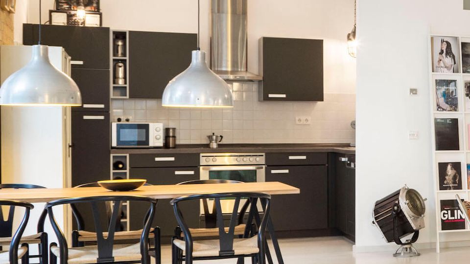 Open-plan modern kitchen and dining area with matte black cabinets, stainless steel range hood, and white tiled backsplash. A long wooden table with black chairs sits under two large silver pendant lights, with high ceilings and a white floor in a minimalist loft-style space.