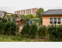 Außenbereich mit einer Schaukel aus Holz, einem Zaun mit grünen Sträuchern und einem kleinen Gartenhaus. Im Hintergrund ein flacheres Wohngebäude mit Dachziegeln und ein mehrstöckiger Wohnblock, umgeben von Bäumen.