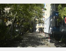 Eine schattige Gasse mit Kopfsteinpflaster, gesäumt von Bäumen und Wohnhäusern. Im Hintergrund ist ein mehrstöckiges Gebäude mit großen Fenstern und einer roten Backsteinfassade zu sehen.