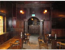 Holzgetäfelter Innenraum einer rustikalen Bar mit schummriger Beleuchtung. Im Vordergrund stehen Holztische und -stühle, während im Hintergrund eine kleine Theke mit Barhockern durch einen gemauerten Rundbogen sichtbar ist.