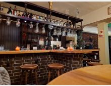 Gemütliche Bar mit Theke aus Holz und Ziegelsteinen, dekoriert mit Kerzen, kleinen Pflanzen und Gläsern, die über Kopf hängen. Im Hintergrund befinden sich Holzregale mit Flaschen und ein Weihnachtsbaum.