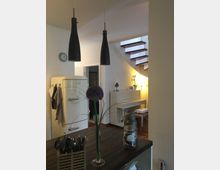 Moderne Innenansicht eines Wohnraums mit offener Küche. Im Vordergrund befindet sich eine Kücheninsel mit dunkler Holzarbeitsplatte, darüber zwei schwarze Hängeleuchten; im Hintergrund ein weißes Klavier, eine freistehende Treppe mit Holzstufen und ein alter Kühlschrank im Retrodesign.