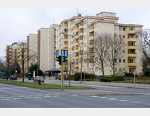 Mehrstöckige Wohngebäude mit hellen Fassaden und gelben Balkonen entlang einer Straße. Im Vordergrund befindet sich eine U-Bahn-Station mit einem markanten gelben "U"-Schild, umgeben von Bäumen, Gehwegen und parkenden Autos.