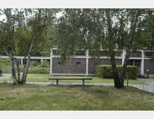 Außenansicht einer modernen Backstein-Gebäudeanlage mit großen Fenstern, umgeben von einer Grünfläche mit Wiese, Birken und Büschen; im Vordergrund steht eine Betonbank.