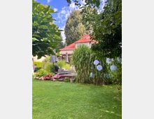 Blick in einen üppig bepflanzten Garten mit Rasenfläche, Holzterrasse und Sitzmöbeln samt Sonnenschirmen. Im Hintergrund steht ein Wohnhaus mit rotem Ziegeldach, umgeben von hohen Bäumen und dichtem Grün mit Blumenbeeten und hängenden Papierlaternen.