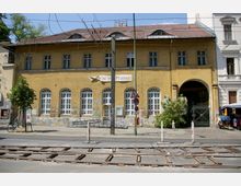 Straßenansicht eines historischen gelben Gebäudes mit Rundbogenfenstern und abblätternder Fassade. Im Vordergrund befinden sich Straßenbahnschienen, Verkehrsschilder und ein Baum, rechts ist ein Torbogen mit schmiedeeisernem Gitter zu sehen.