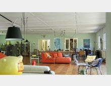 Großer, hell gestalteter Wohnraum mit Holzboden und einer weißen Decke. Im Vordergrund befinden sich orangefarbene Sofas, ein Couchtisch und Stühle; im Hintergrund sind Regale mit Büchern, ein langer Esstisch und diverse Wanddekorationen zu sehen.