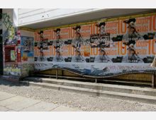 Fassade eines Gebäudes mit Pflastersteinweg davor; die Wände sind mit bunten Plakaten, darunter Werbung für ein Event, und Graffiti bedeckt. Rechts führt ein gewelltes Geländer eine kleine Treppe hinunter, links befindet sich eine überwiegend rote Anzeigetafel.