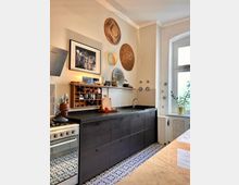 Moderne Küche mit schwarzen Unterschränken, schwarzer Arbeitsplatte und einem Gasherd mit Edelstahlfront. An der cremefarbenen Wand hängen dekorative Körbe und ein gerahmtes Foto, während ein großes Fenster mit natürlichem Licht und Pflanzen die Szene ergänzt.