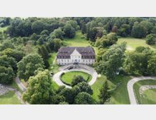 Luftaufnahme eines historischen Gebäudes mit heller Fassade und rotem Dach, umgeben von einem großen, bewaldeten Park. Vor dem Gebäude befindet sich eine kreisförmige Zufahrt, die von grünen Wiesen und Bäumen eingerahmt ist.