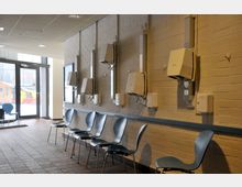 Ein Wartebereich mit einer Reihe von blauen Kunststoffstühlen entlang einer hellen Ziegelwand, an der mehrere Trockengeräte und Seifenspender angebracht sind. Im Hintergrund ist eine Glastür mit Blick auf den Außenbereich sichtbar.