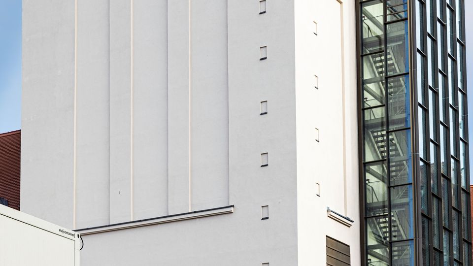 Außenansicht eines weißen Theatergebäudes mit glatter Fassade und rotem „THEATER“-Schriftzug auf dem Dach. Rechts schließt ein moderner, verglaster Anbau mit sichtbarem Treppenhaus und Metallrahmen an.