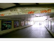 Unterführung mit gelblichen Fliesenwänden, an denen gerahmte Poster angebracht sind. Die Decke zeigt Spuren von Verfall und Graffiti, der Ausgang im Hintergrund ist hell und führt ins Freie, wo einige Personen erkennbar sind.