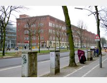 Straßenansicht mit einem großen Backsteingebäude im Hintergrund, das mehrere Stockwerke und rechteckige Fenster hat. Im Vordergrund gibt es Bäume, einige Verkehrsschilder, Fahrradständer und Stromkästen; eine Straße mit parkenden Autos und Geschäften im Erdgeschoss des Gebäudes ist sichtbar.