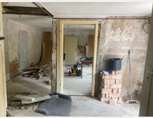 Ein renovierungsbedürftiger Innenraum mit freiliegenden Backsteinwänden, abblätterndem Putz und Werkzeugen. Im Vordergrund liegen Baumaterialien und ein Eimer steht auf einem Stapel Ziegelsteine.