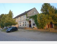 Ansicht eines Dorfgasthofs mit historischem Gebäude, das teilweise mit Efeu bewachsen ist. Vor dem Haus stehen Bäume, ein schwarzes Auto ist auf einer unbefestigten Fläche geparkt, und im Vordergrund befindet sich ein Kopfsteinpflasterweg.