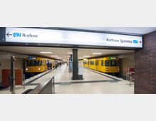 U-Bahn-Station mit zwei parallel liegenden Gleisen, auf denen gelbe U-Bahn-Züge stehen. Beschilderungen weisen auf die Linienrichtung der U7 mit den Zielen "Rudow" und "Rathaus Spandau" hin. Der Boden ist gefliest, und die Beleuchtung ist hell.