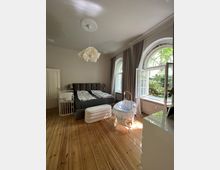 Ein helles Schlafzimmer mit Holzdielenboden, einem Doppelbett, einer Wiege und einem Babybett. Rechts befinden sich große, halbhohe Fenster mit Rundbögen, die viel Tageslicht hereinlassen, daneben sind lange graue Vorhänge angebracht.