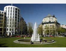 Öffentlicher Platz mit einem zentralen Springbrunnen, umgeben von Grünflächen, Sitzbänken und Menschen, die die Umgebung genießen. Im Hintergrund moderne, mehrstöckige Gebäude und Bäume, die den Platz einrahmen.
