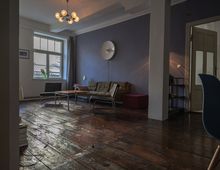 Wohnzimmer mit dunklem Holzboden, einer Wand in violetter Farbgebung, modernen Möbeln wie einem Sofa und Couchtischen sowie einer großen Fensterfront mit Blick auf ein Dach. An der Decke hängt eine Lampe mit mehreren kleinen Leuchtkugeln, und im Hintergrund führt eine Tür in einen weiteren Raum.