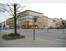 Straßenkreuzung in einer Stadt mit mehrstöckigen historischen Gebäuden im Hintergrund, darunter ein Eckgebäude mit roter Ladenfront. Straßenschilder mit den Namen „Stromstraße“ und „Alt-Moabit“, Fußgänger, Autos, sowie eine U-Bahn-Markierung und ein Baum im Vordergrund.