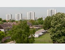 Städtische Landschaft mit Wohngebäuden: Im Vordergrund sind ein kleiner Parkplatz, grüne Bäume und Wiesen zu sehen, dahinter befinden sich Fachwerkhäuser mit tiefen Dächern. Im Hintergrund ragen mehrere hohe, weiße Plattenbauten empor, umgeben von weiteren Bäumen und Vegetation.