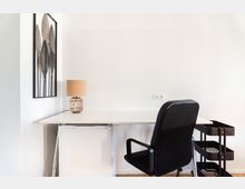 Minimalistischer Arbeitsbereich mit weißem Schreibtisch, schwarzem Bürostuhl und schwarzem Rollwagen. An der Wand hängt ein metallisches Kunstwerk mit Blättermuster, und auf dem Tisch steht eine Tischlampe mit einem Korbgestell.