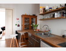 Helle Küche mit Holzarbeitsplatten, moderner Spüle und offenen Wandregalen, die mit Gläsern und Küchenutensilien bestückt sind. Im Hintergrund steht ein hölzerner Vitrinenschrank, und an der Wand hängen kleine bunte Bilder; im Vordergrund ein Bartresen mit zwei Hockern.