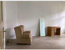 Ein leerer Raum mit weißen Wänden und einem beigen Sessel auf der linken Seite, einem schmalen, auf dem Boden stehenden Spiegel in der Mitte und einem kleinen Holzschreibtisch mit Schubladen auf der rechten Seite. Der Boden besteht aus glattem, unbehandeltem Material.