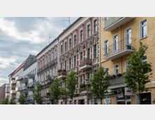 Straßenansicht mit mehrstöckigen Altbau- und Wohngebäuden, teils mit verzierten Fassaden und Balkonen. Im Erdgeschoss befinden sich Geschäfte, darunter ein Pizzaladen, und Bäume säumen den Gehweg.