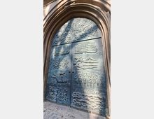 Große bronzene Doppeltür in einem hohen, steinernen Spitzbogenportal, mit reliefartigen Szenen und Figuren auf den Türflügeln. Im oberen Bereich ist ein Baumrelief zu sehen; die Oberfläche wirkt patiniert und wird von schrägem Sonnenlicht teilweise beleuchtet.