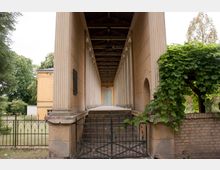 Klassischer Säulengang mit hohen, verzierten Säulen und einem hölzernen Dach, das zu einer geschlossenen grünen Tür am Ende führt, flankiert von einem schmiedeeisernen Tor und umgeben von einer parkähnlichen Landschaft. Rechts ist eine Mauer mit rankenden Pflanzen zu sehen.