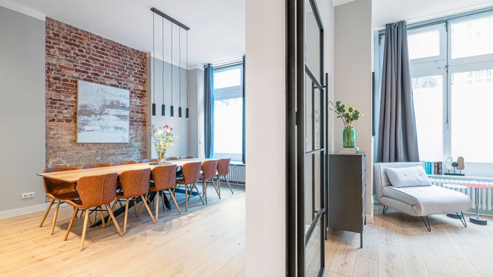 Heller, moderner Wohn- und Essbereich mit freiliegender Backsteinwand, langem Holzesstisch mit mehreren Stühlen und schlichten Hängeleuchten. Rechts daneben eine Sitzecke mit grauer Chaiselongue vor großen Fenstern mit Vorhängen, getrennt durch eine schwarze Glastür im Industriestil.