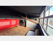 Industrieartiger Innenraum mit Holzfußboden, roter Wandfläche und schwarzer Stahlkonstruktion sowie einer schrägen Rampe zu einer erhöhten Ebene. Rechts verläuft eine Reihe großer, alter Sprossenfenster mit Blick auf die umliegenden Dächer.