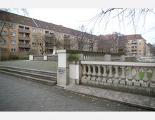 Wohnhäuser mit mehreren Balkonen im Hintergrund, davor eine gepflasterte Fläche mit steinernen Treppenstufen und einem Geländer aus Balustern. Kahle Bäume und ein kleines begrüntes Areal sind sichtbar, die Umgebung wirkt städtisch.