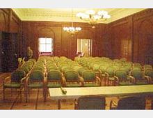 Ein Konferenzraum mit einer Reihe grün gepolsterter Stühle in mehreren geraden Linien, einer weißen Tischreihe im Vordergrund und Holzvertäfelungen an den Wänden. Eine Tür und ein Fenster lassen Tageslicht in den Raum, während an der Decke ein großer Kronleuchter hängt.