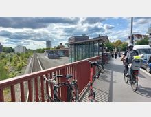 Ein Bahnhof mit Bahngleisen links, flankiert von Grünflächen und Gebäuden im Hintergrund. Rechts eine Fußgänger- und Fahrradbrücke mit abgestellten Fahrrädern und einem E-Scooter, sowie Passanten und ein Radfahrer im Vordergrund.