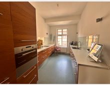 Lange, schmale Küche mit modernen Holzschränken und weißen Arbeitsflächen. Auf der linken Seite befinden sich ein eingebauter Ofen und Schubladen, auf der rechten eine Arbeitsfläche mit Spüle, Wasserhahn und Computer; im Hintergrund ist ein Fenster, das Tageslicht hereinlässt.