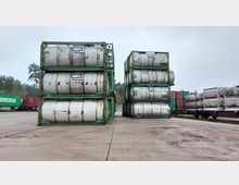 Gestapelte ISO-Tankcontainer auf einem Lagerplatz, umgeben von weiteren Behältern und Containern auf Güterzügen; im Hintergrund ist ein Wald unter grauem Himmel sichtbar.