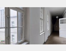 Ein heller Flur mit weißen Wänden und Holzboden, beleuchtet durch mehrere große Sprossenfenster auf der linken Seite. Rechts ist eine geöffnete weiße Tür mit Glaseinsatz zu sehen, und im Hintergrund befindet sich ein dunkler Vorhang.