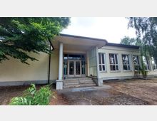 Eingangsbereich eines einstöckigen Gebäudes mit heller Fassade, großen rechteckigen Fenstern und einem Vordach, das von zwei Säulen gestützt wird. Der Eingangsbereich ist über eine kleine Treppe erreichbar, die von Bäumen und Sträuchern umgeben ist.