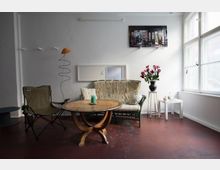 Minimalistisch eingerichtetes Wohnzimmer mit einem runden Holztisch in der Mitte, einem gemusterten Sofa, einem beigegrünen Klappstuhl und einem weißen Beistelltisch mit Blumenstrauß. An der linken Wand hängen Haken, und ein großes Fenster rechts lässt viel Tageslicht herein.