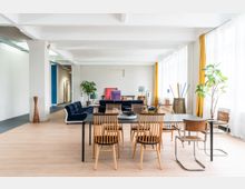 Offener, heller Raum in einem modernen Büro- oder Wohnbereich mit großen Fenstern, weißen Vorhängen und gelben Akzenten. Holzböden, ein Esstisch mit Holzstühlen, eine blaue Samtcouch, Zimmerpflanzen und bunte Dekorationen prägen das Interieur.