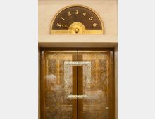 Ein goldener, kunstvoll verzierter Fahrstuhl mit floralen Mustern auf den Türscheiben; darüber eine halbrunde Etagenanzeige mit goldenen Zahlen von E bis 6 und einem Zeiger in klassischem Design.