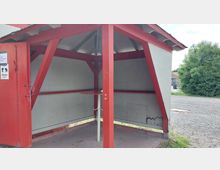 Kleine, offene Wetterschutzhütte mit rot gestrichenen Holzstreben und einem Satteldach. Im Inneren befindet sich ein runder Stehtisch, und die Umgebung zeigt einen gepflasterten Boden sowie grüne Büsche im Hintergrund.