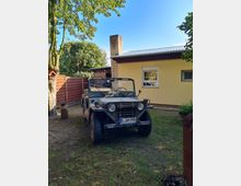 Ein offenes, olivgrünes Militärfahrzeug steht auf einer Rasenfläche vor einem gelben Gebäude mit einem Schornstein und Satteldach. Im Hintergrund sind ein Holzzaun, Bäume und blauer Himmel zu sehen.