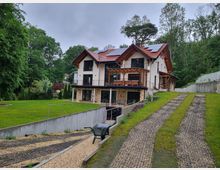 Zweistöckiges modernes Haus im Landhausstil mit weißen Wänden, dunklen Holzelementen und rotem Ziegeldach, umgeben von einem großen, gepflegten Garten mit Bäumen im Hintergrund. Eine Einfahrt aus Kies führt leicht geneigt zum Gebäude, während Sonnenkollektoren auf dem Dach sichtbar sind.