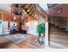 Treppenhaus in einem historischen Gebäude mit einer geschwungenen Holztreppe, verzierten Geländern und rosafarbenen Wänden, dekoriert mit Geweihen. Im Vordergrund steht eine grüne Zimmerpflanze auf einem kleinen Tisch, und an der Wand hängen historische Informationen, die von zwei Personen betrachtet werden.